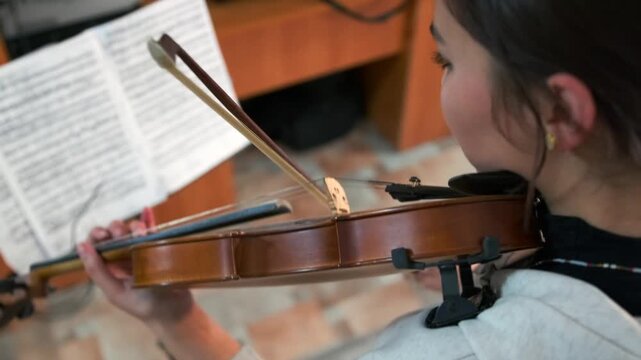 Young female violin student nervously sight-reading sheet music during an exam or audition. Tense posture and reflect the emotional pressure and focus required in classical training. Side view.