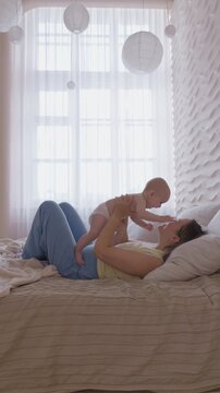 mother lifting baby above chest on bed. morning exercise and play mom with infant with bright window light. cheerful movement and eye contact. happy domestic maternal routine. cozy bonding