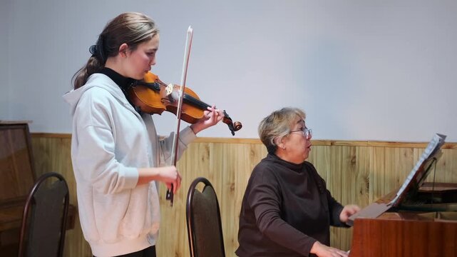 Teenage girl playing violin with piano accompaniment during a classical music lesson. Elderly teacher playing piano and watching the student attentively. Rehearsal in a music school classroom.