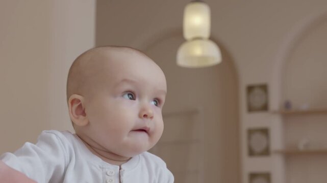 Caucasian baby sitting on bed and putting fingers in mouth. Thoughtful wide eyes and gentle chewing motion. Neutral tones of the nursery and soft authentic lighting. 