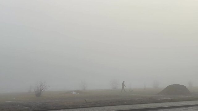 solitary walker in dense fog across barren roadside field, distant silhouette moving past lamp posts and muddy mounds muted gray tones, chilly dawn atmosphere, quiet commute and isolation captured