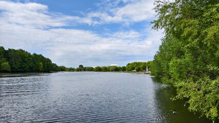 Willows grow on the grassy shores of the lake. There is a park with a beach on the shore, and buildings behind it. There are ripples on the water and buoys installed. Sunny spring weather and blue sky © Balser