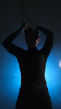 Vertical video of a skilled young man's silhouette as he performs a flow art routine with spinning nunchaku against a smoky blue background.