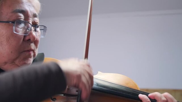 Elderly Asian woman playing violin with focused expression. Close-up of fingers and bow on the strings -  shot indoors in warm soft lighting. Senior woman playing the violin during practice session