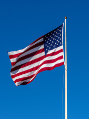 Fototapeta premium American Flag Flown Against Clear Blue Sky, Waving Proudly On Tall Flagpole In Bright Sunlight