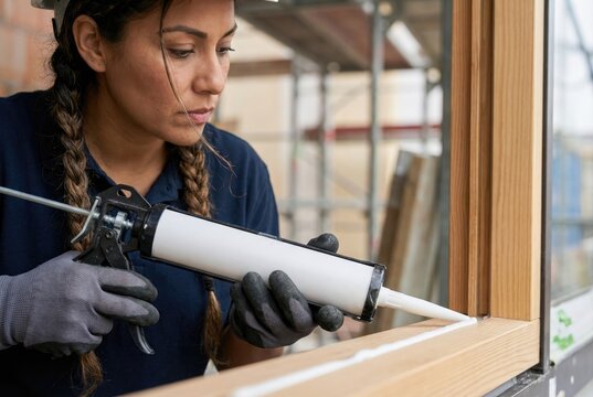 A female construction worker uses a caulking gun to apply a bead of white silicone sealant to a wooden window frame.