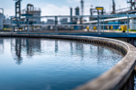 Industrial wastewater treatment facility with water settling in a circular clarifier tank under an overcast sky showing metal railings and blurred processing equipme