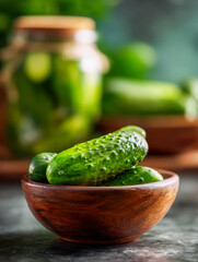 Fresh green cucumbers in a wooden bowl with blurred jar of pickles in the background on rustic surface in natural light setting