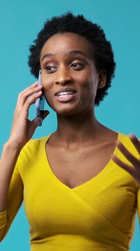 Vertical video of a happy young African American woman talking on her smartphone and smiling, isolated on a blue background.