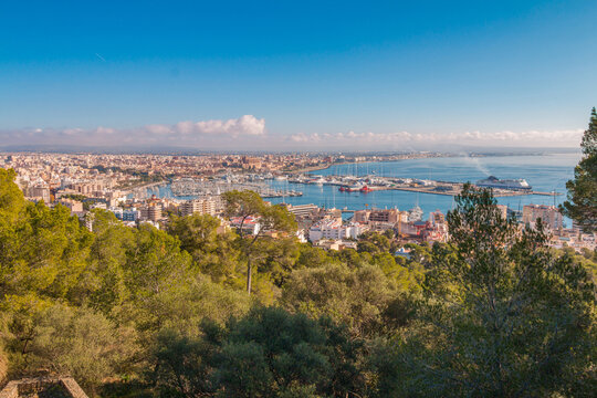 View from Carrer Castell de Bellver over Palma de Mallorca on a sunny day, panorama of the harbor, rooftops, blue sea and mountains in the background, Mediterranean atmosphere and warm, bright light.