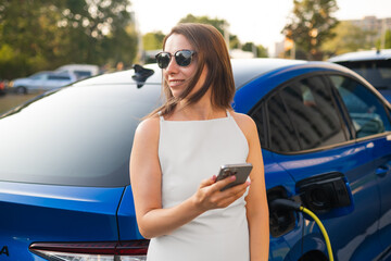Naklejka premium Woman in sunglasses stands next to her electric car, checking her phone as it charges