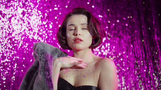 Close-up of a woman's face, with glitter on her cheekbones and shoulders, leaning on her gray fur coat, against a pink-magenta glitter background. 4K video.