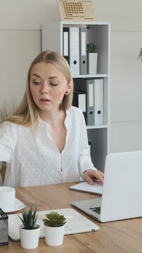 A young businesswoman works on a laptop and takes notes in a notebook while sitting at her desk in a modern office. Vertical video.