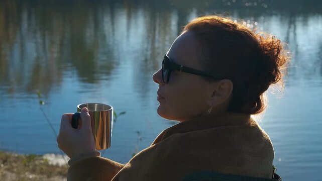 Woman with sunglasses drinking hot tea outdoors. Relaxed woman in sunglasses sitting by a calm lake, enjoying a hot steaming drink from a metal camping mug while contemplating the serene natura