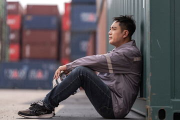 A tired logistics worker in a gray uniform sits on the ground at a busy shipping container yard,...