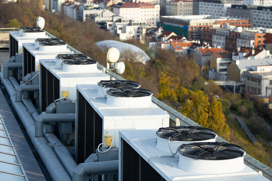 Rooftop view of multiple air conditioning units with fans and pipes, overlooking a cityscape with buildings and trees in the background during daylight
