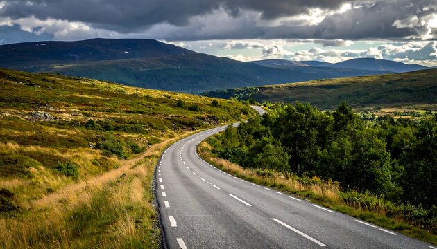 Asphalt road curves through rolling green hills under a dramatic sky, with glimpses of distant peaks