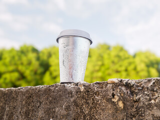 ​Silver metallic beverage cup mockup with condensation droplets on a stone wall. Blurred nature...