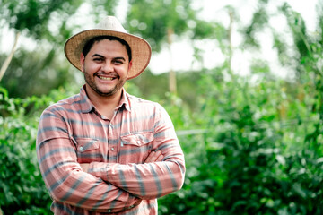 Portrait of a smiling male farmer wearing a straw hat and plaid shirt standing with arms crossed in...