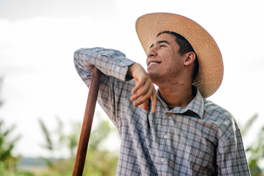 Happy young male farmer leaning on a wooden tool handle in a rural field, wearing a straw hat and plaid shirt.