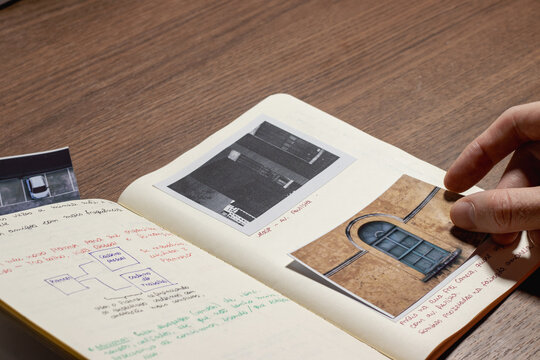 Person's hand placing a printed photograph into a personal journal or notebook with handwritten notes and other pictures on a wooden desk, symbolizing creative process, planning, and memory.