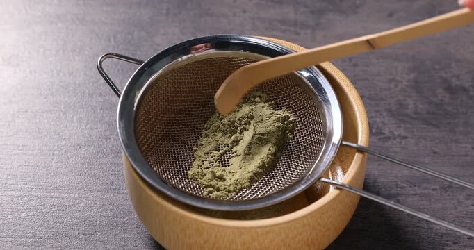 Woman sifting green matcha powder into ceramic bowl at gray textured table, closeup