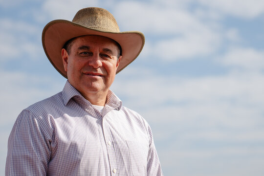Portrait of a smiling mature man wearing a cowboy hat and a checkered shirt standing outdoors against a bright blue sky