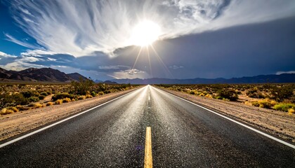 A straight, black asphalt road leads towards the bright sun in a blue sky. Rolling hills and desert vegetation surround it