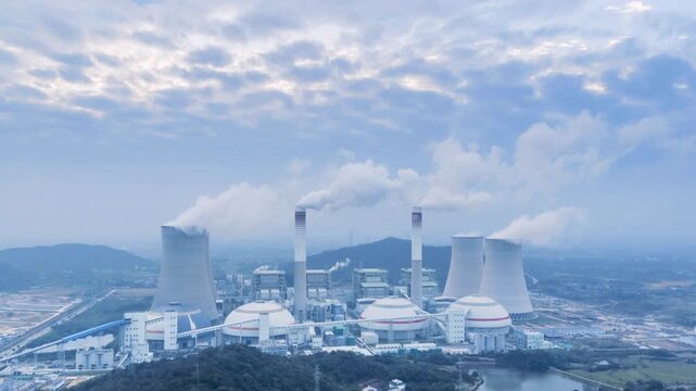 Aerial time-lapse of a coal-fired power plant at cloudy early morning, industrial landscape of cooling towers and chimneys emitting steam.