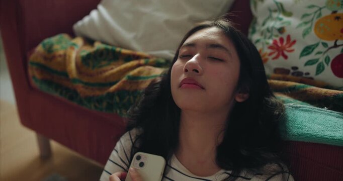 teenage girl reclines on the floor by a rust sofa, eyes closed, relaxing, holding a white smartphone. Patterned cushions and a green gold throw sit nearby in soft natural daylight.