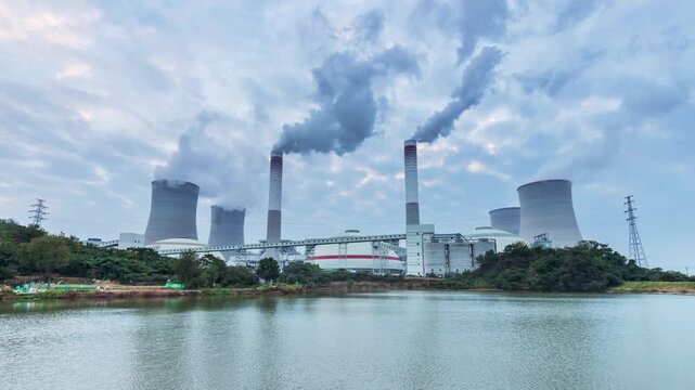 Early-morning time-lapse of a coal-fired power plant, cooling towers and smokestacks emitting steam, reflected in calm water under cloudy skies.