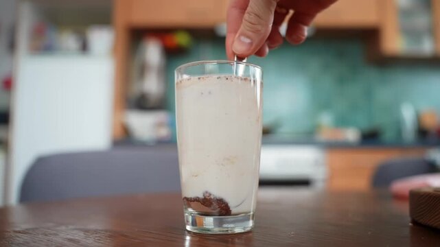 Hand stirs a tall glass of chocolate milk with cocoa sediment at the bottom on a wooden table, soft-focus home kitchen background. Cozy drink preparation close-up.