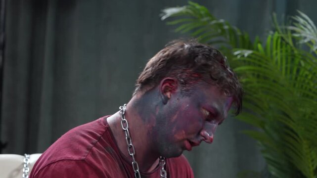 A man with purple body paint and red shirt sits indoors with a heavy silver chain around his neck, his face and expression partially smeared and theatrical in style