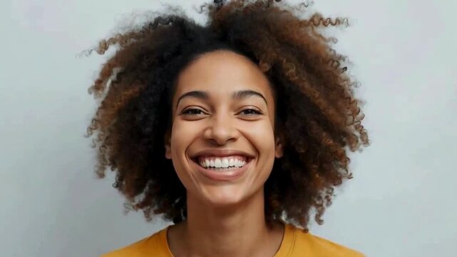 A joyful young woman with curly hair smiles brightly, showcasing happiness and confidence against a simple backdrop.	happy video
