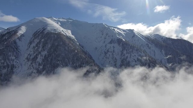 Aerial view of sunny sky above clouds drifting through snowy mountain ranges and deep alpine valleys. Winter light highlights dramatic peaks and layered clouds across high elevation terrain.