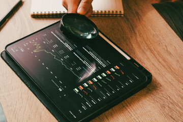 Close up of financial expert using magnifying glass to audit stock market charts on digital tablet, analyzing investment data details.