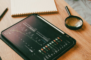 Professional investment workspace with stock market trading charts on tablet and magnifying glass on wooden desk.
