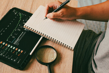 Businessman writing financial strategy in notebook with stock market charts on tablet, concept of investment planning and data analysis.