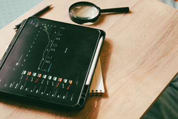 Professional investment workspace with stock market trading charts on tablet, magnifying glass, and stylus on wooden desk.