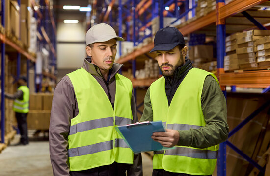 Workers in warehouse review inventory on clipboard. Shelves of boxes surround them as discussion on tracking steps and delivery accuracy for fulfillment. Concept shows smart logistics flow.