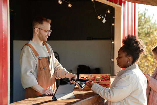 Caucasian young adult man processing payment with card reader for Black woman buying strawberries at outdoor market stall, both focused on transaction, digital tablet visible