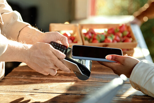 Caucasian man holding payment terminal while young Black woman making contactless payment with smartphone at outdoor market, fresh strawberries visible in background, hands only