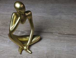 Golden Abstract Sculpture of Contemplative Human Figure. A modern golden sculpture depicting a stylized seated human figure in a thoughtful pose. Crafted with smooth, elongated limbs and a minimalist  © Framed Essence