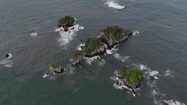 A detailed high-angle drone shot capturing the powerful Atlantic waves hitting a small rocky island near the shore of Limbe
