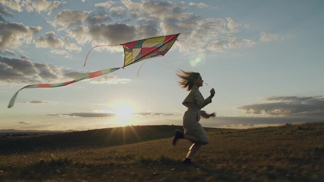 Young woman runs across grassy field flying colorful kite during golden hour sunset. Scene evokes freedom joy and summer vibes. Perfect for lifestyle and travel commercials.