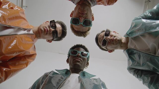 Four stylish men in shiny jackets and sunglasses look down at the camera from a low angle. A cool and confident fashion shot suitable for youth culture or lifestyle ads.