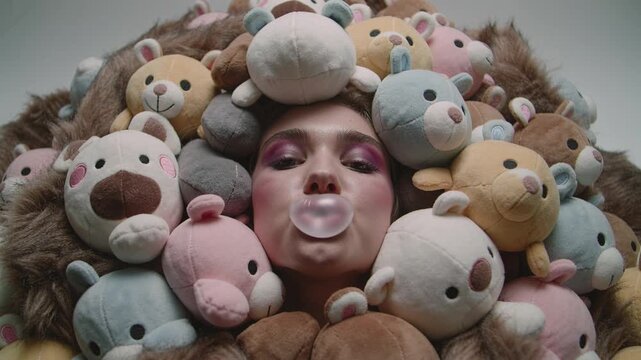 Young woman face surrounded by soft teddy bears blowing pink bubble gum. Playful and cute atmosphere for fashion or lifestyle concepts. Ideal for creative advertising and youth themes.