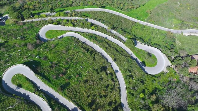 AERIAL TOP DOWN: Winding mountainside road with sharp switchbacks and vehicles navigating the bends. Pale asphalt snakes through lush green shrubbery and trees on west Mediterranean coast of Sicily.