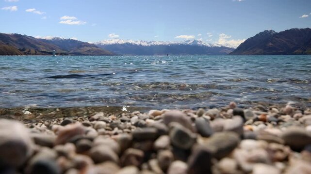 Static close focus shot on waves crashing on a pebble beach of Lake Hawea. Senic snowcapped mountrain in background.
