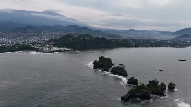 A wide drone vista capturing the atmospheric beauty of a Cameroonian bay, with dark volcanic outcrops scattered across the calm water under a misty sky